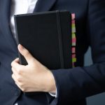 A businesswoman in a suit holds a planner with colorful tabs in an office setting.