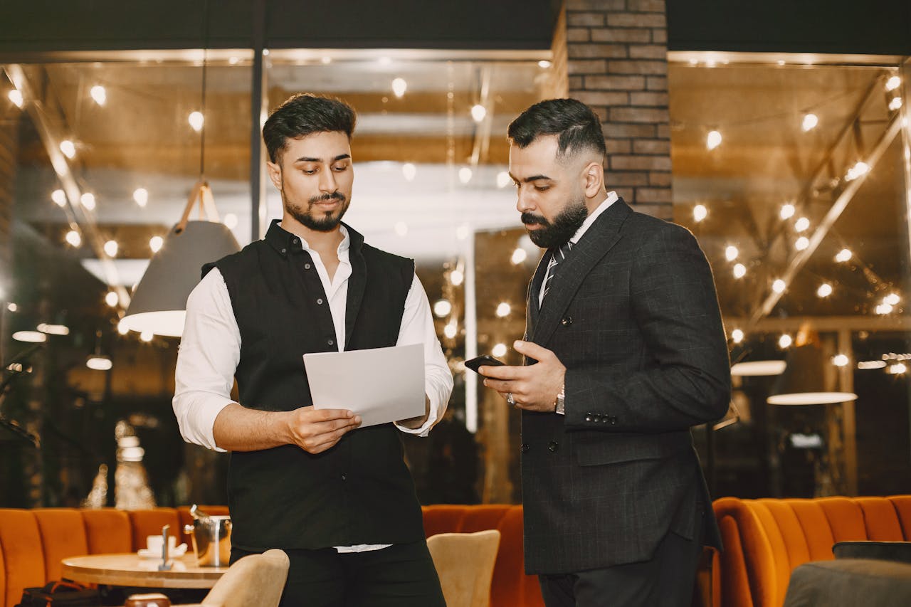 Two businessmen reviewing documents and smartphone in a restaurant setting with warm lighting.